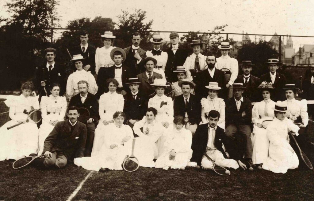 Ffotograff du a gwyn o dynion a menywod yn eistedd i'r gamera gyda'i racedi tenis 