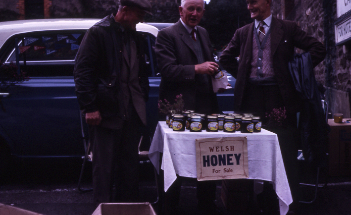 Llun lliw o dri dyn ar stondin fechan gydag arwydd 'Welsh HONEY For Sale', ac arddangosfa o jariau mêl.
