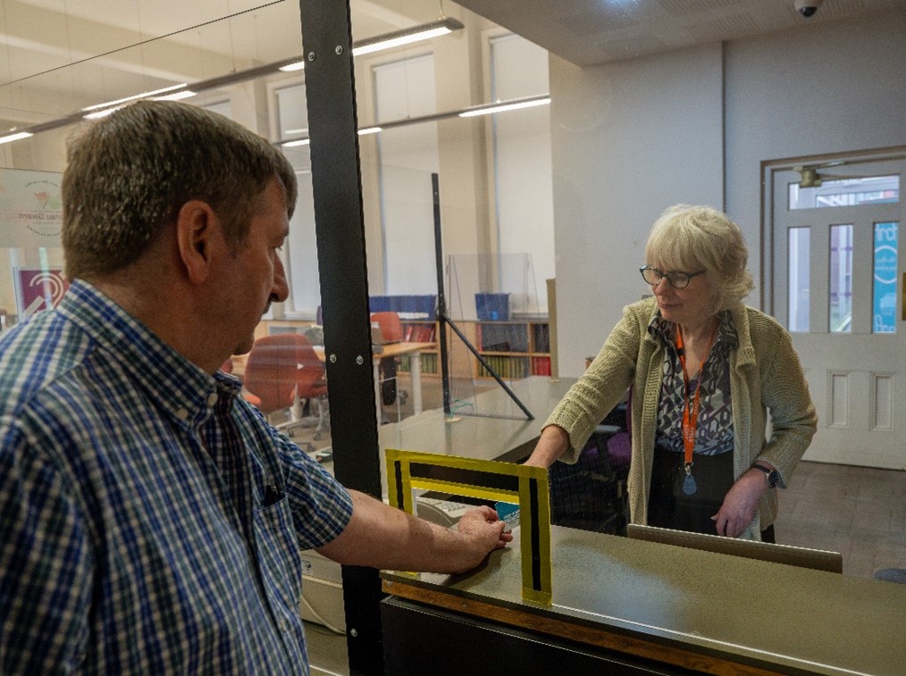 Mae aelod o staff yn y dderbynfa y tu ôl i sgrin cownter ac yn derbyn cerdyn gan aelod o'r cyhoedd trwy fwlch bach yn y sgrin. Mae ffiniau'r bwlch yn y sgrin wedi'u marcio â thâp.